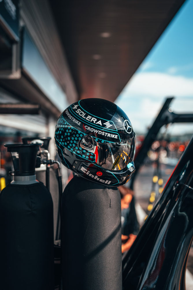 Un casco del equipo Mercedes-AMG PETRONAS Formula One Team, con los logotipos de los sponsors claramente visibles —entre ellos Einhell, CrowdStrike y Solera—, está apoyado sobre una garrafa en la calle de boxes. El reflejo en la visera muestra el entorno del garaje.