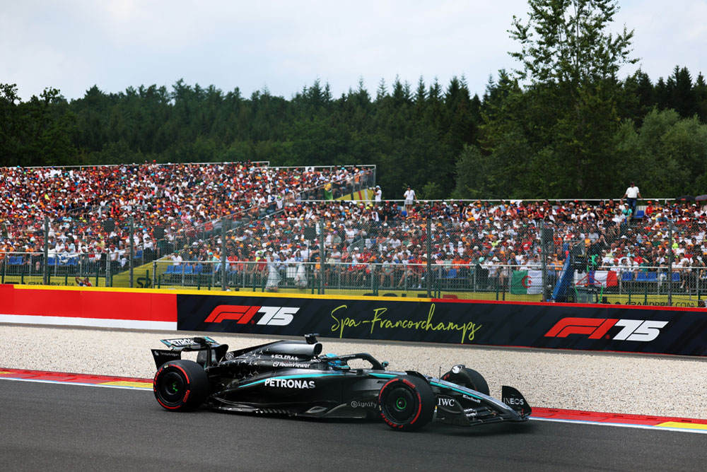 Un auto del equipo Mercedes-AMG PETRONAS Formula One Team corre en el circuito de Spa-Francorchamps. Al fondo, las gradas están repletas de fanáticos que agitan banderas de distintos países. El logo de Spa-Francorchamps es visible en la barrera.