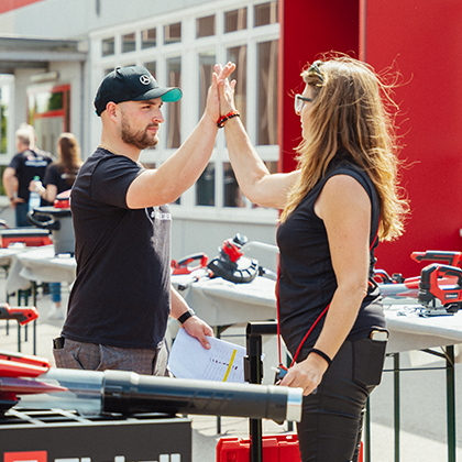 Un hombre y una mujer se chocan las manos en un evento al aire libre de Einhell con exhibición de herramientas, en color.