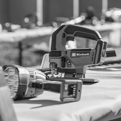 Una sierra caladora inalámbrica Einhell sobre una mesa, con una lámpara inalámbrica al frente, foto en blanco y negro.