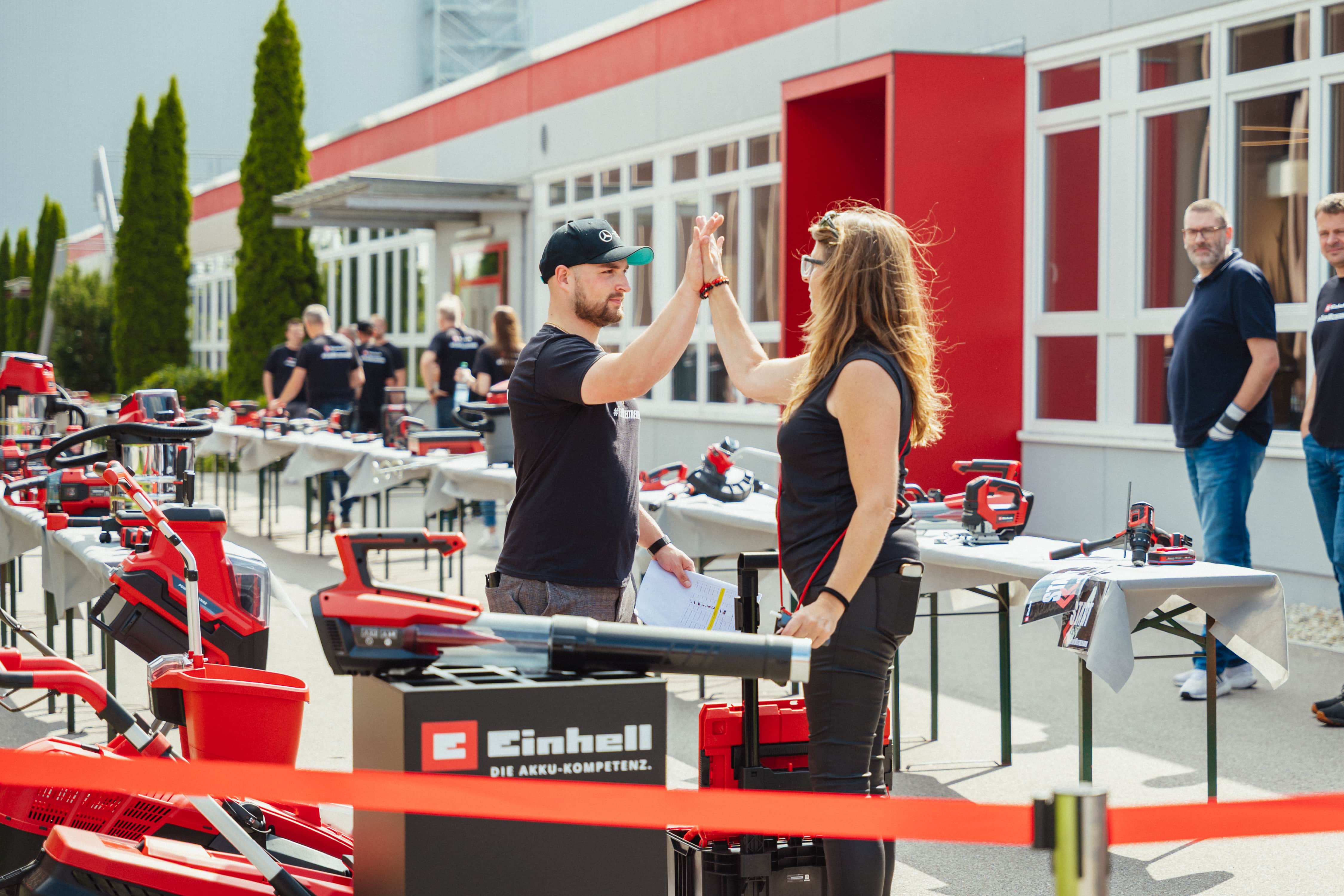 A man and a woman give each other a high five among displayed Einhell tools outdoors.