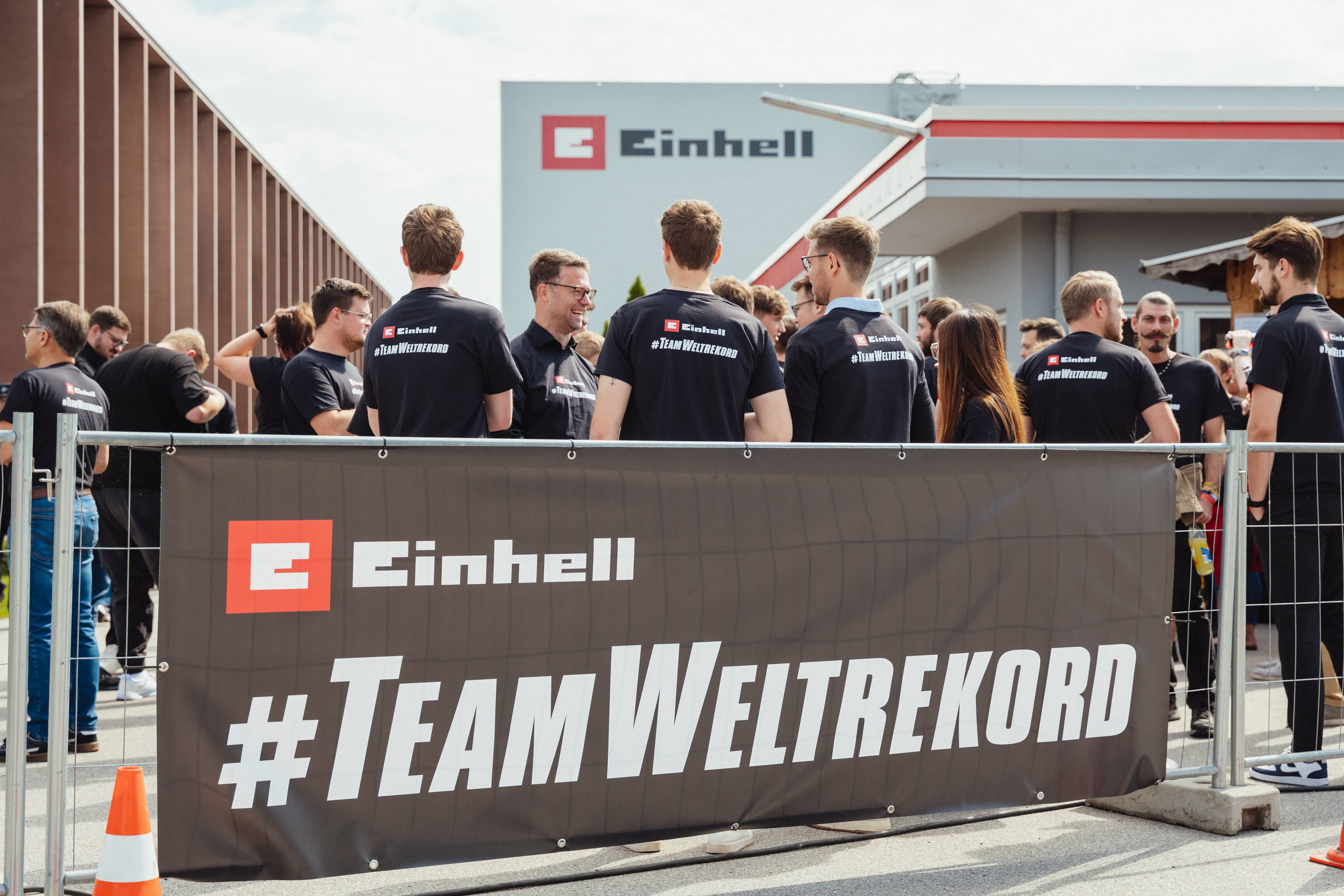 Participants stand behind a fence with a large “Einhell #TeamWeltrekord” banner on the company premises.