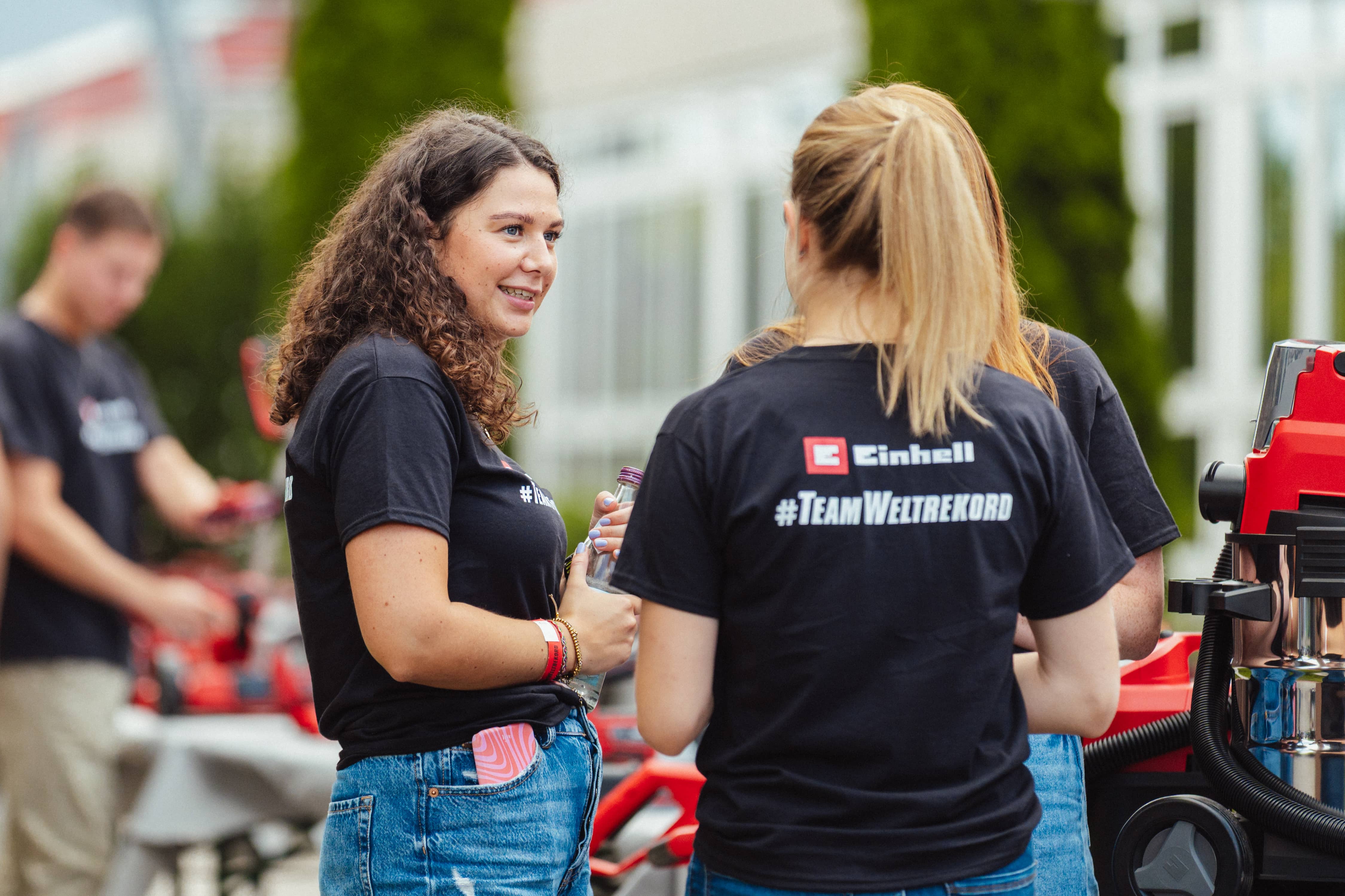 Two young women in "#TeamWeltrekord" shirts talk in front of Einhell tools outdoors.