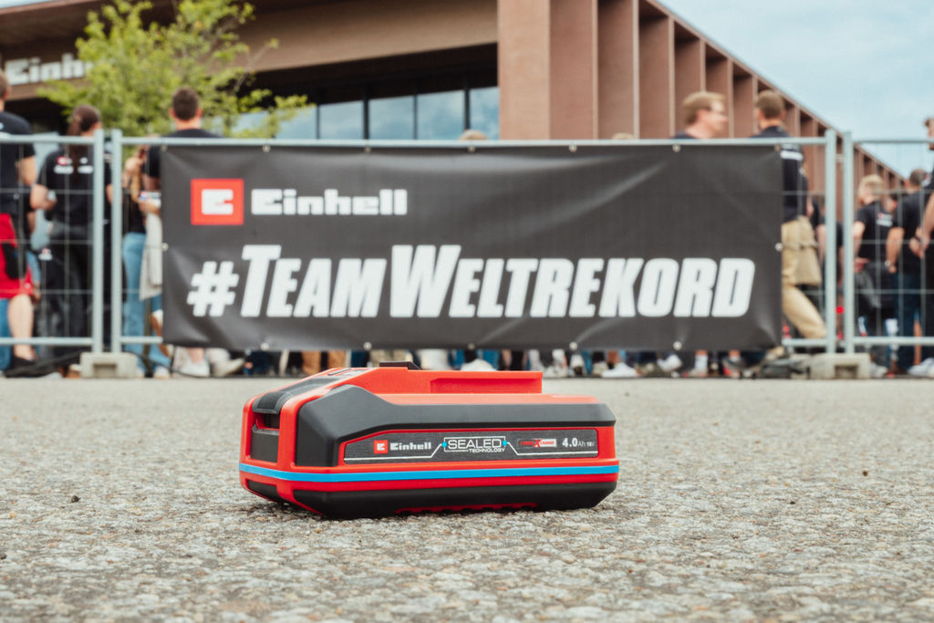 An Einhell SEALED battery Power X-Change 4.0 Ah lies on asphalt, with a "#TeamWeltrekord" banner in the background.
