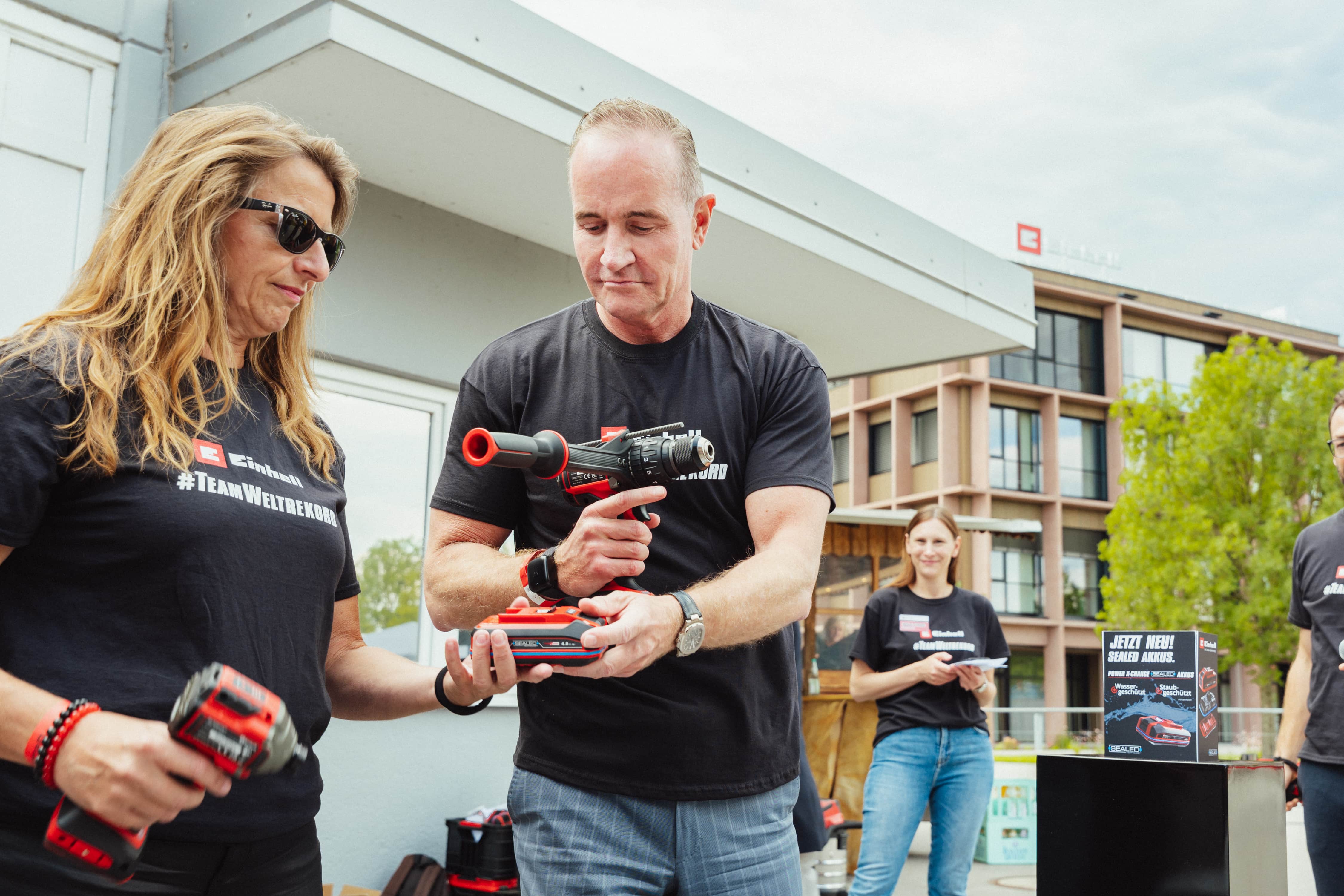 Two people in "#TeamWeltrekord" shirts pass a SEALED battery, one holding a cordless drill.