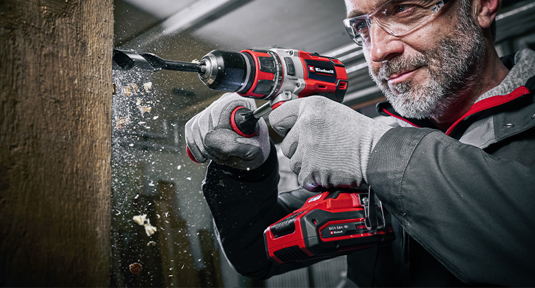 A man drills a hole in a wooden piece with a cordless drill, producing wood shavings.